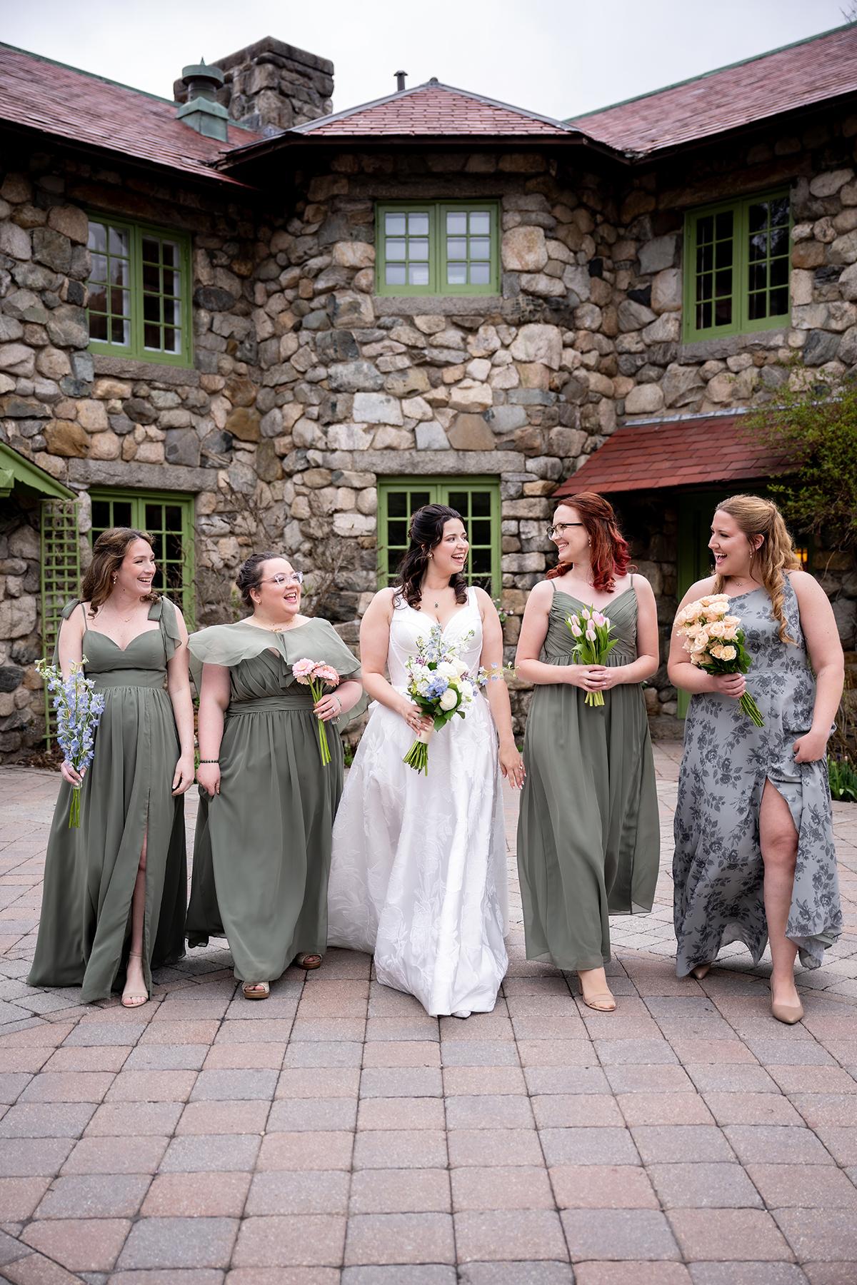 Bride and bridesmaids in dresses, smiling and walking in front of a stone building.