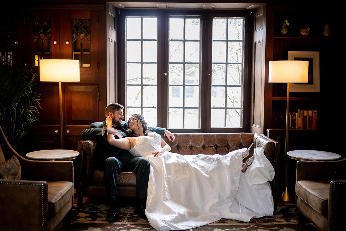 Couple in wedding attire relaxing on a couch, ambiently lit by floor lamps.