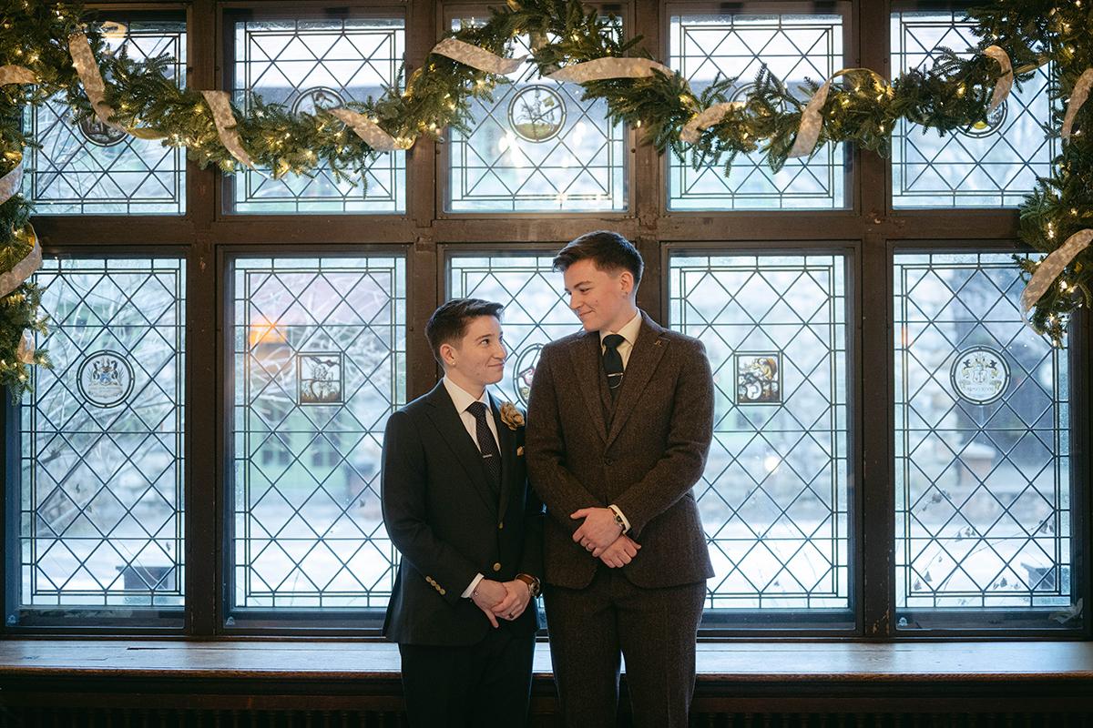 Two people in suits stand smiling under festive garland by large windows.