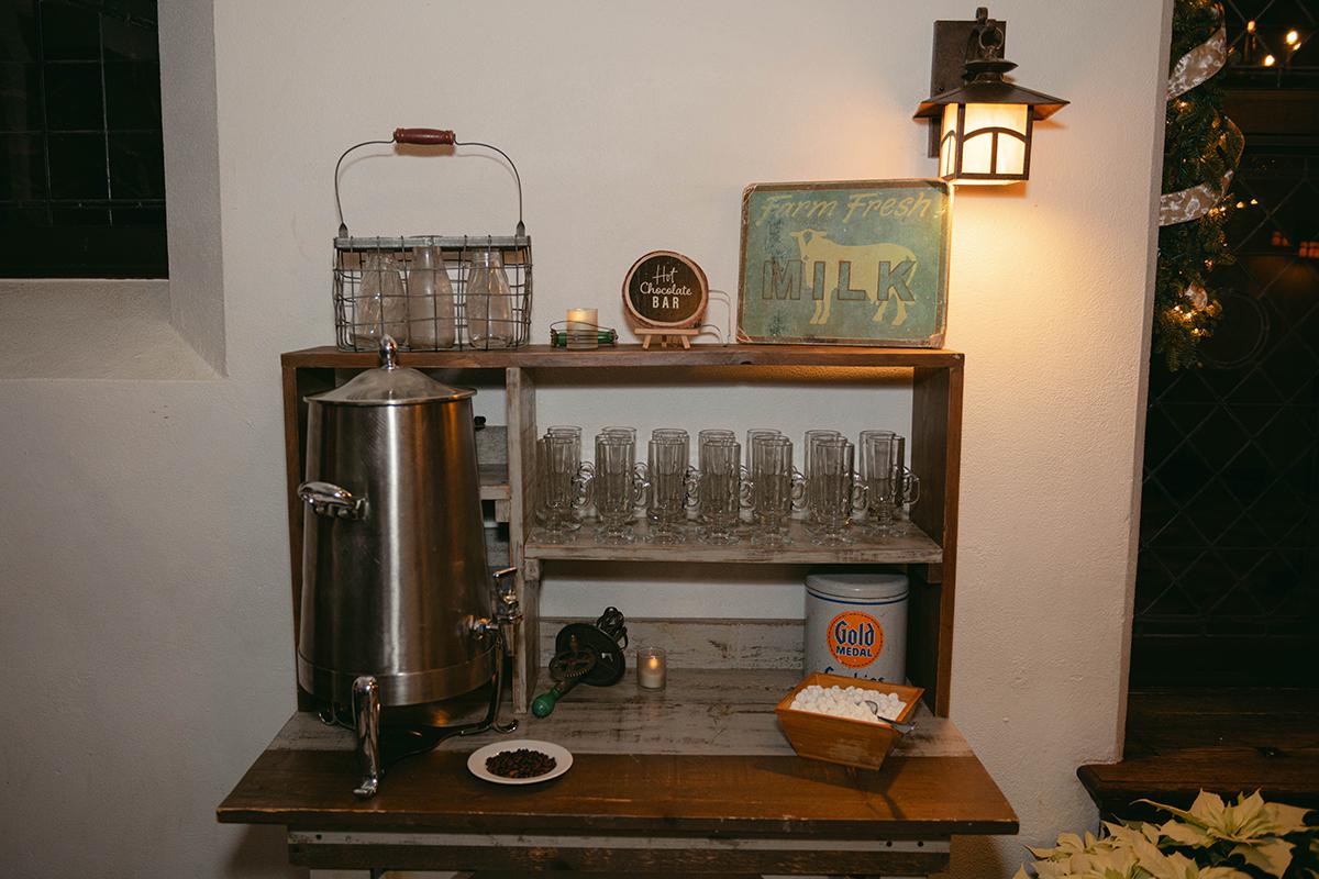Rustic beverage station with metal dispenser and glasses on shelves. Warm lighting.