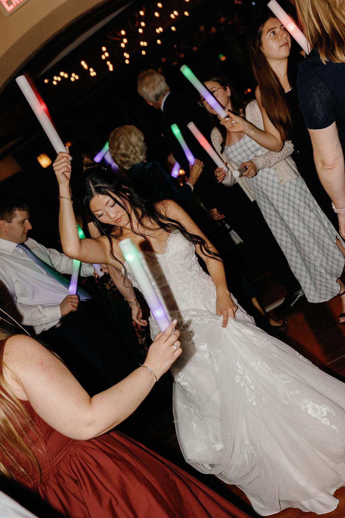 Wedding dance floor, guests holding glowing sticks, bride in white dancing joyfully.