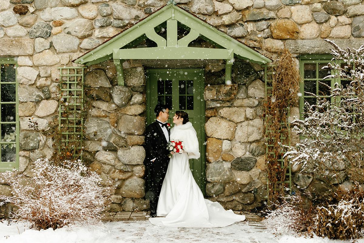Bride and groom in winter attire kissing by a stone cottage entrance.
