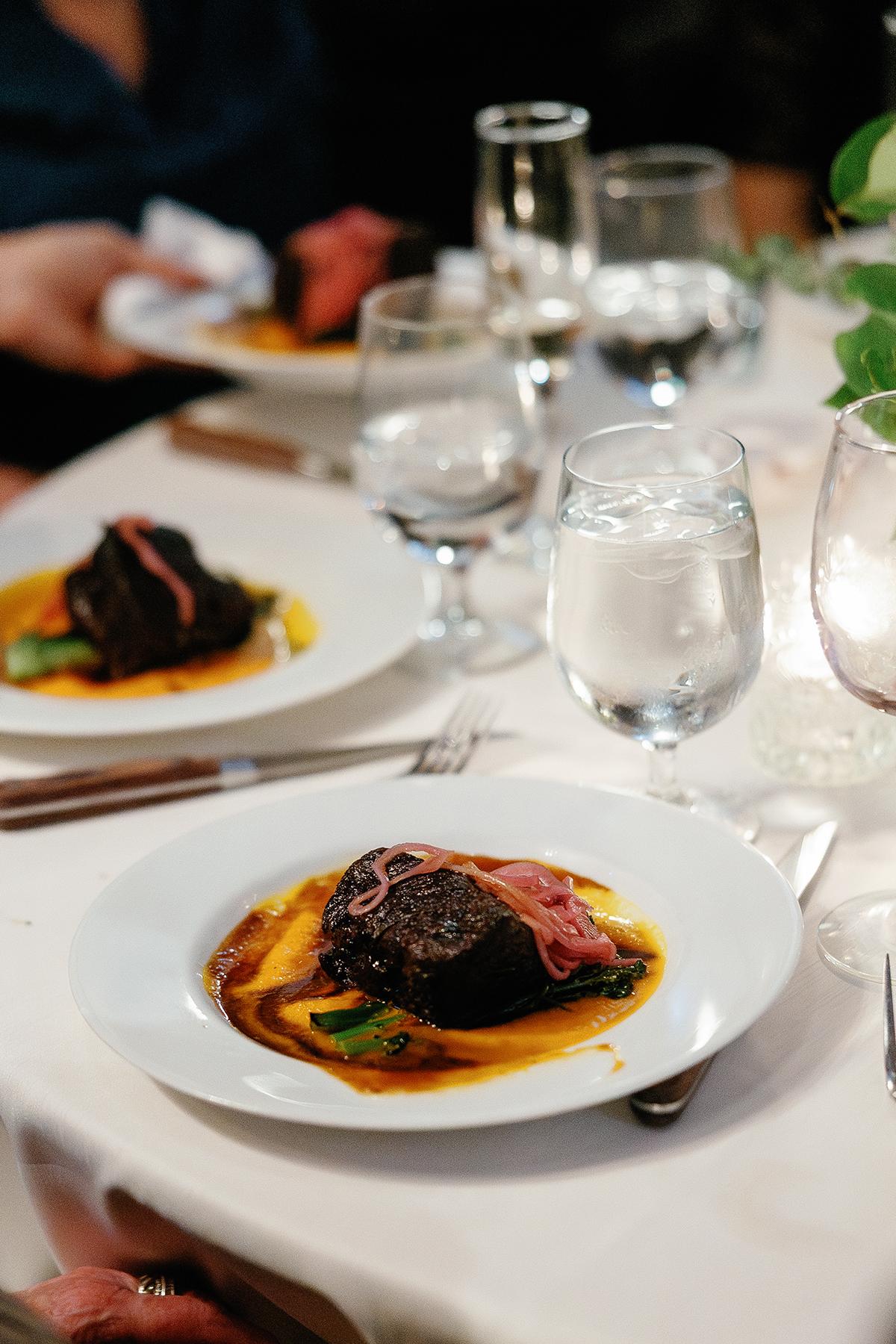 Plated steak with sauce and greens, elegant dining setting.