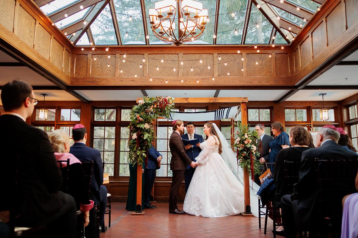 Wedding ceremony with bride and groom under a decorated arch in a sunlit room.