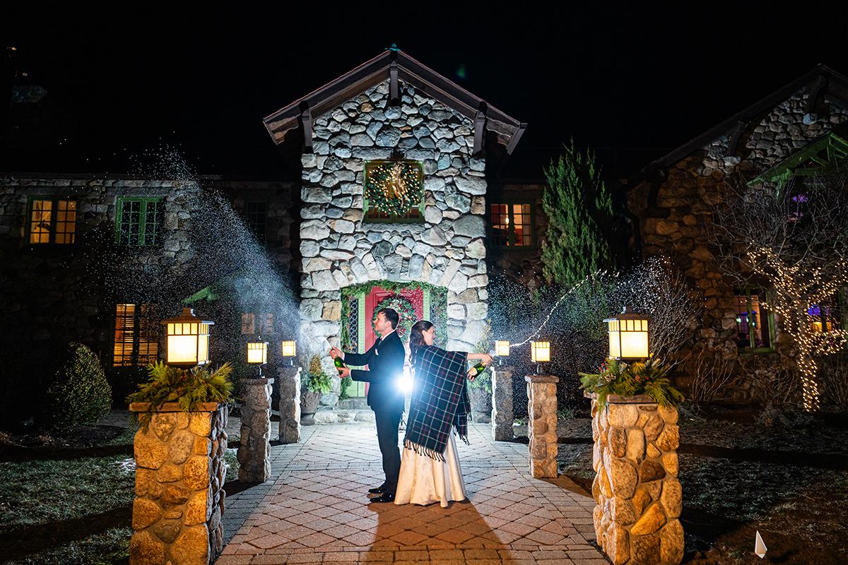 Couple in wedding attire stands illuminated outside stone house at night.