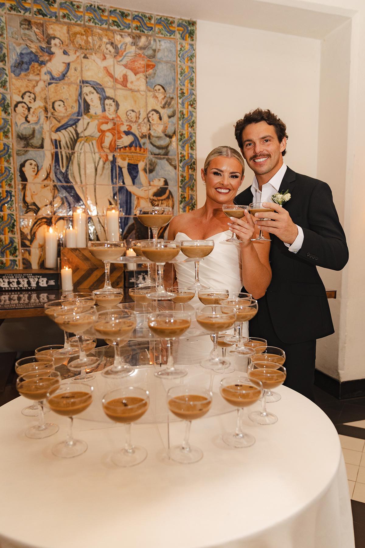 Bride and groom smiling beside a tower of champagne glasses.