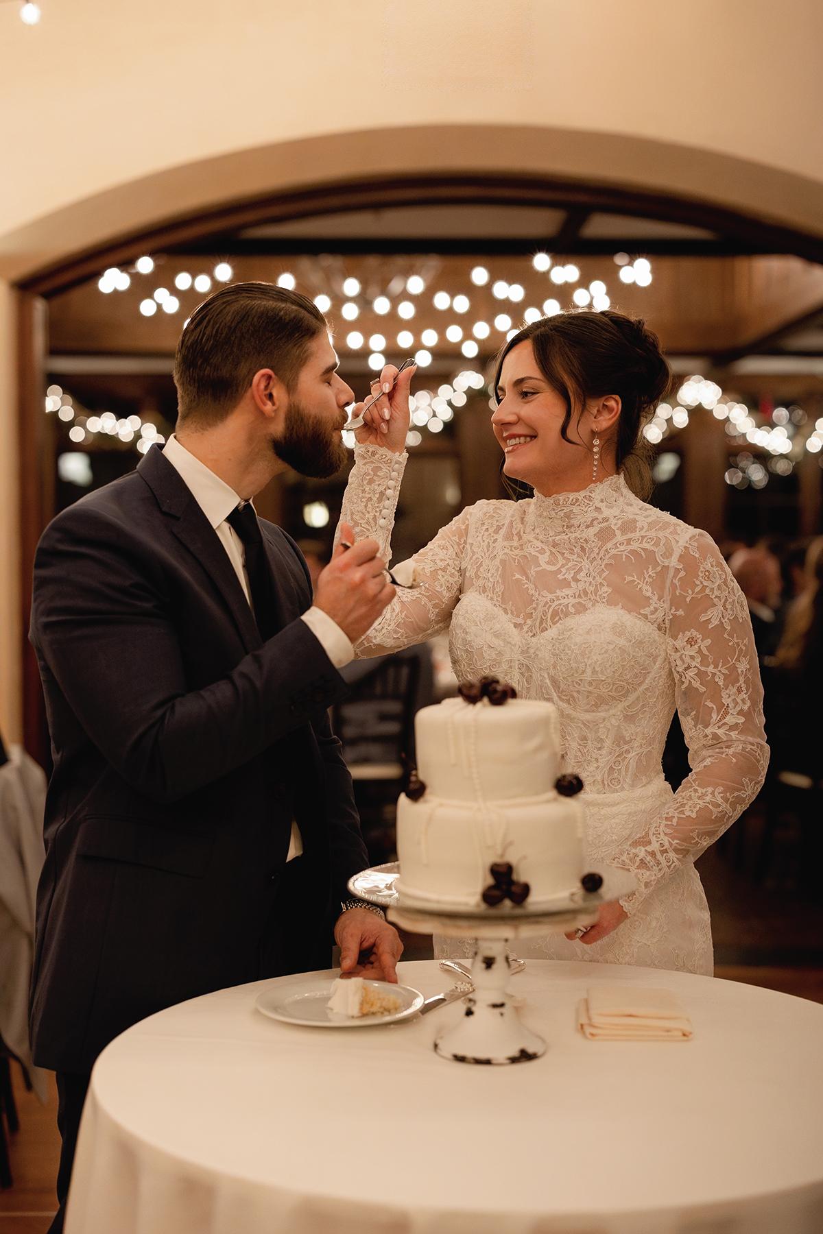 Bride and groom sharing cake, smiling under string lights.
