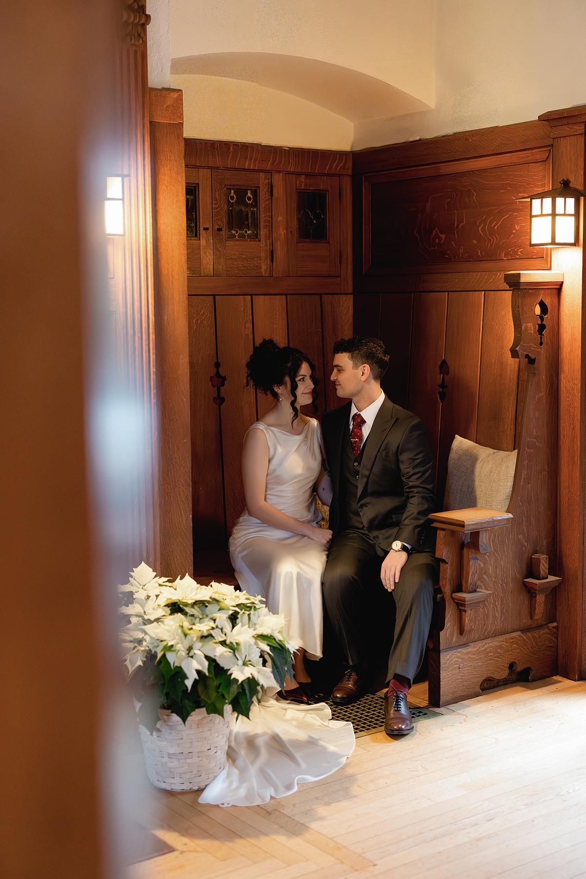 Couple sitting close on a wooden bench, surrounded by warm lighting and a poinsettia plant.
