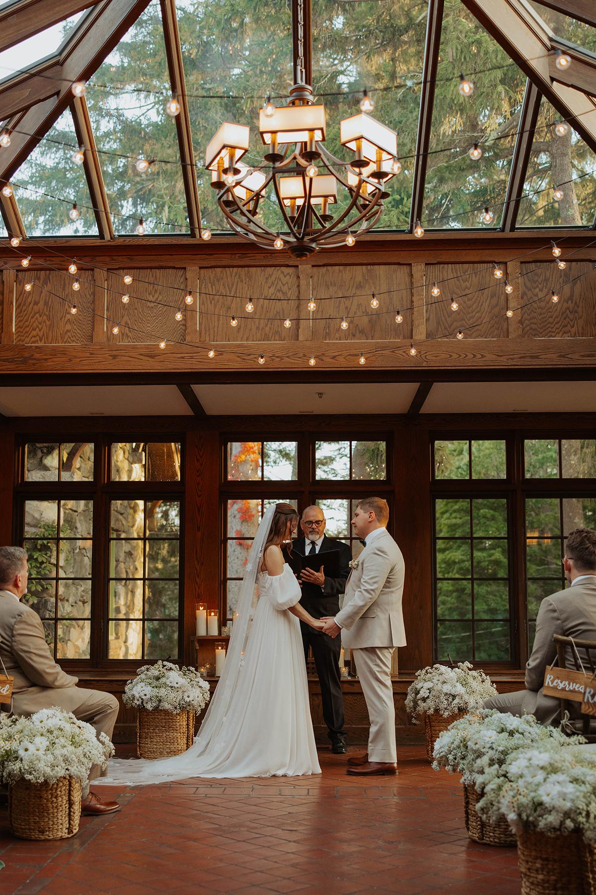 Elegant wedding ceremony in a sunlit room with chandeliers and flowers.