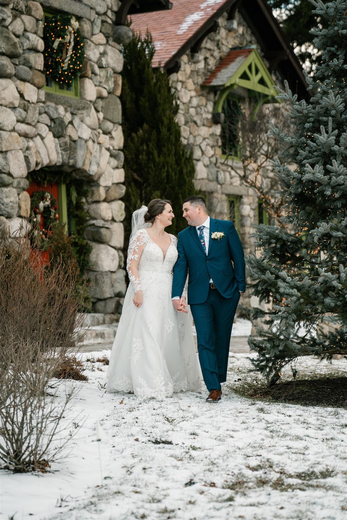 Bride and groom walking on snowy path by a stone building.
