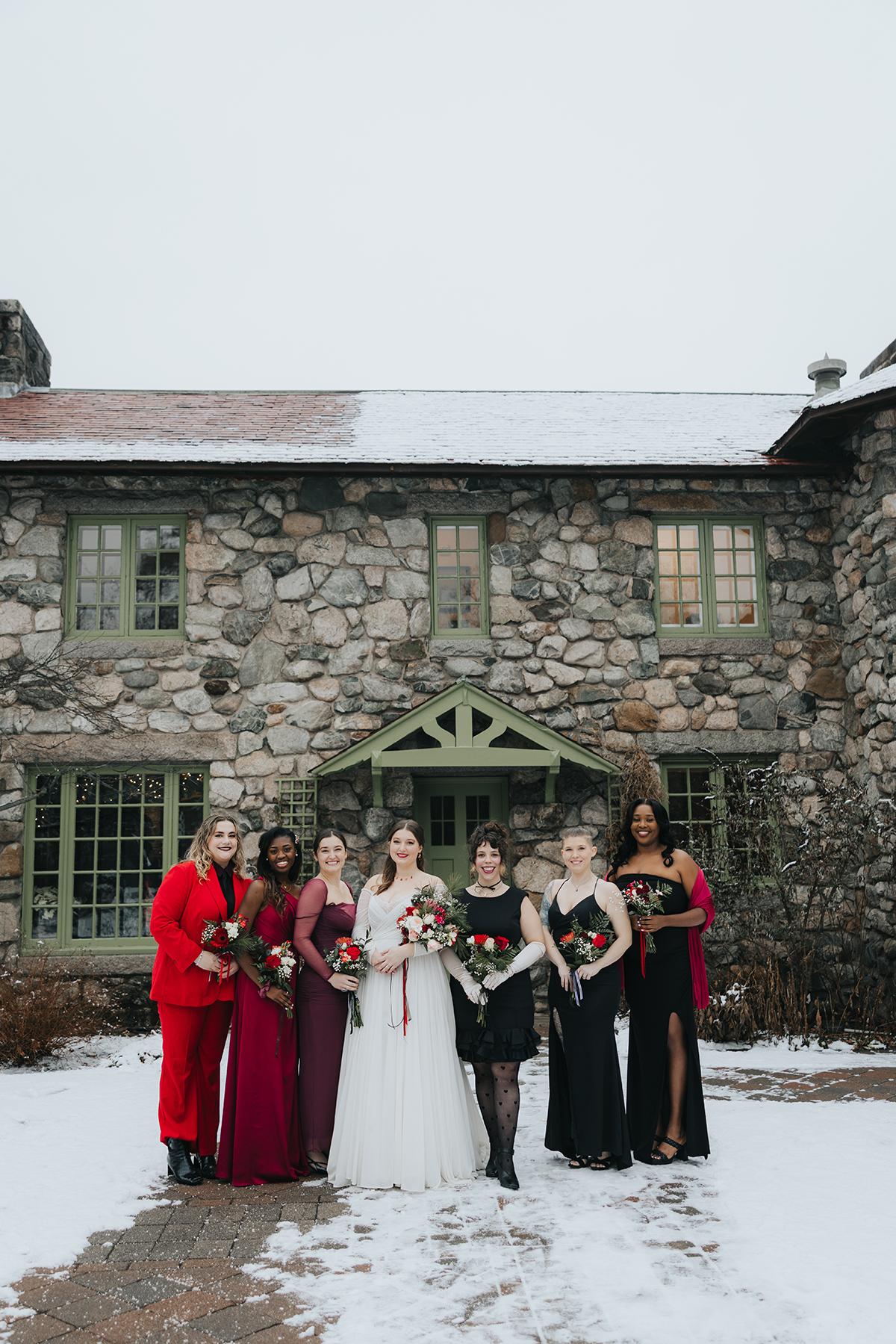 Bridal party in winter attire, posing in front of a stone building with snow on the ground.