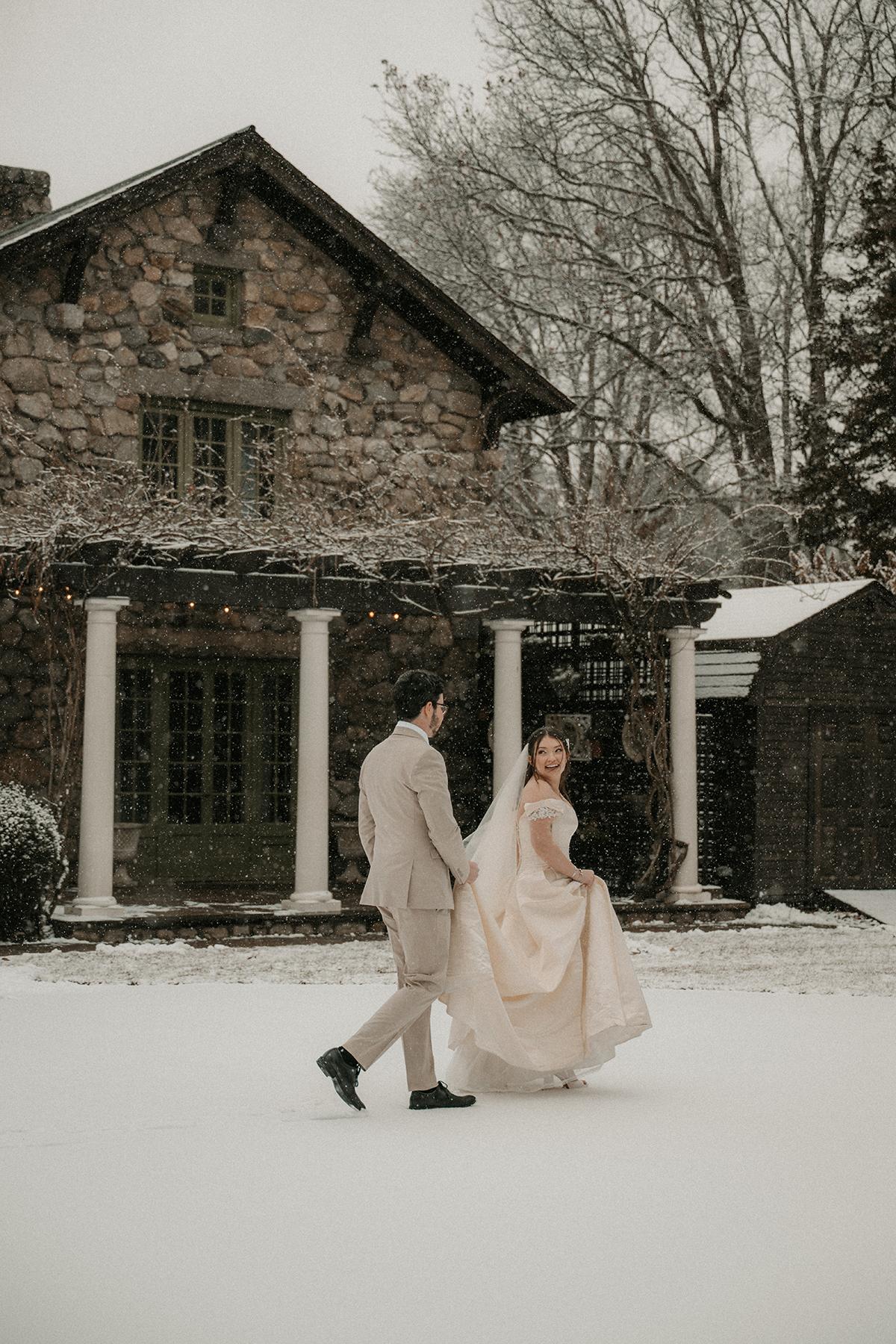 Bride and groom walk through snow beside a stone house.
