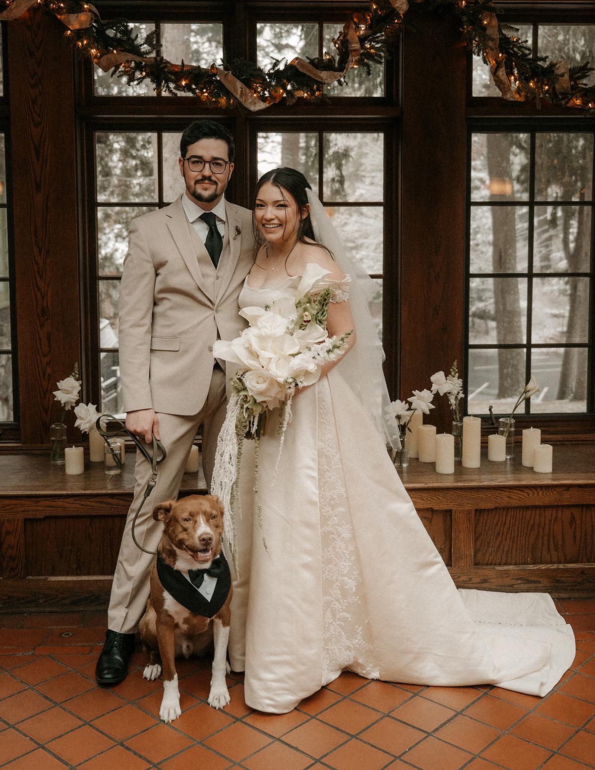 Bride and groom with dog indoors, surrounded by candles and garlands.