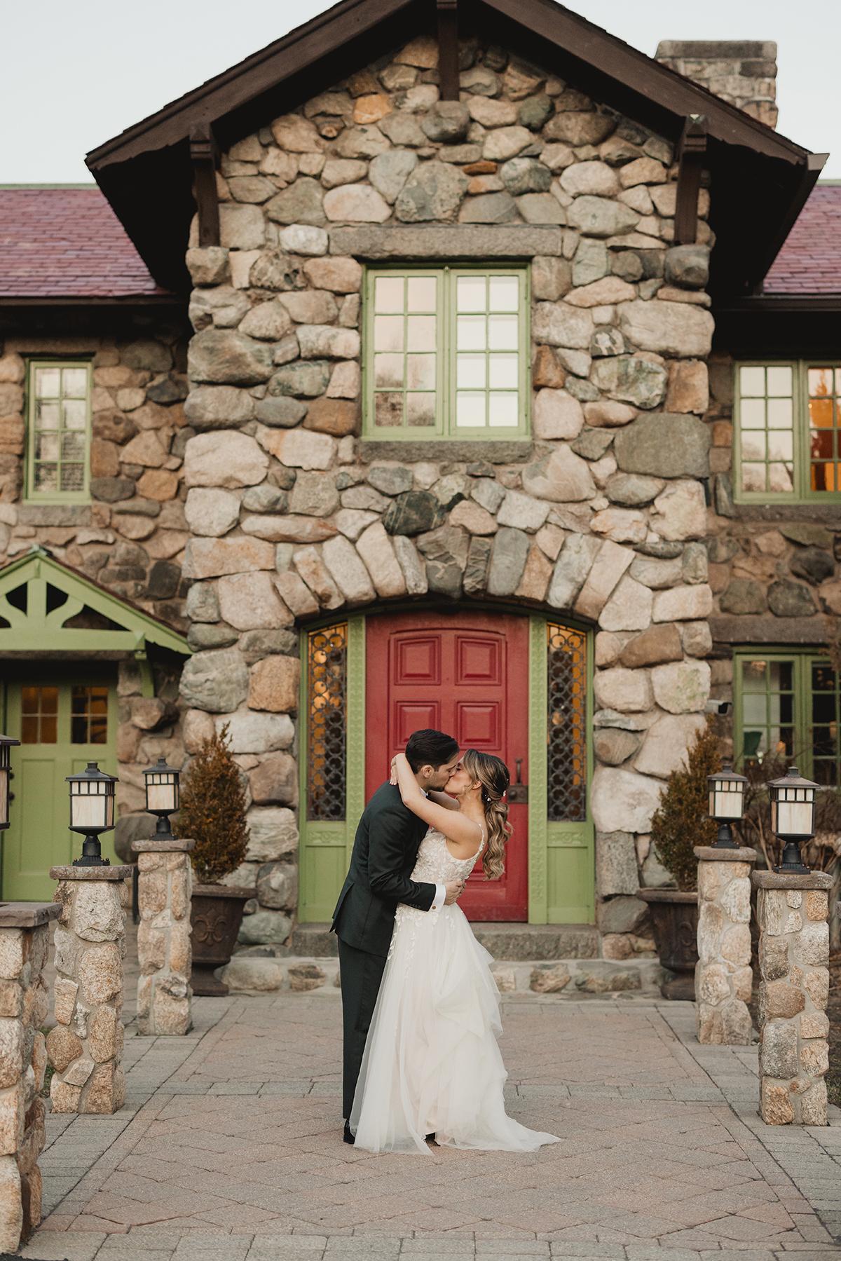 Bride and groom embrace in front of a rustic stone house with a red door.