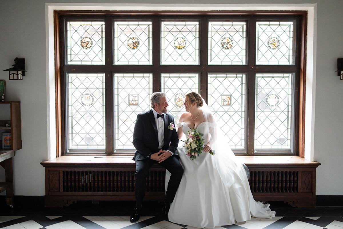 Bride and groom sit smiling by a large ornate window.