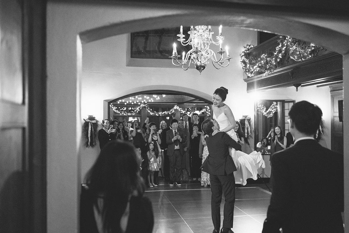 A couple dancing in a decorated hall with onlookers, in black and white.
