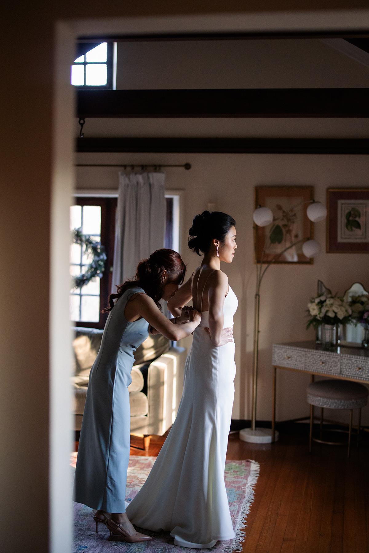 Woman helps bride with dress in softly lit room.