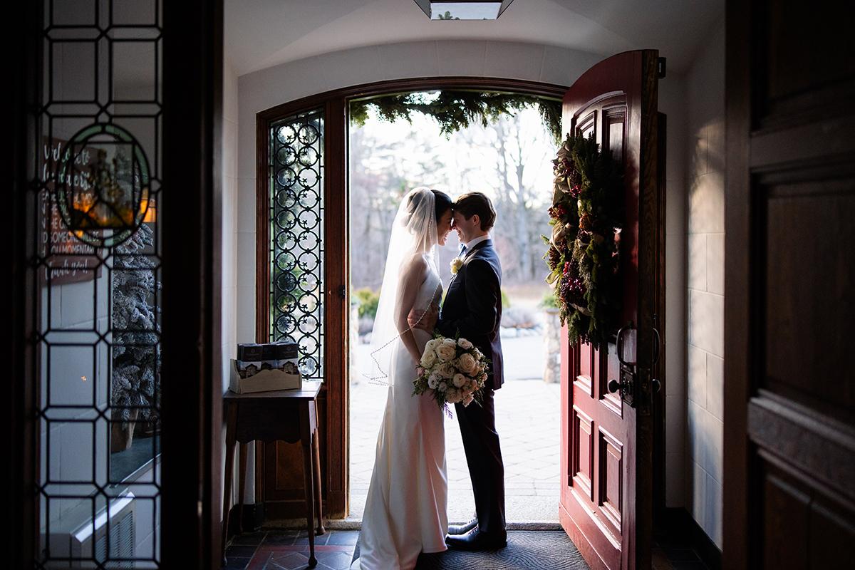 Newlyweds share a kiss in a doorway, sunlight streaming in, bride holding flowers.
