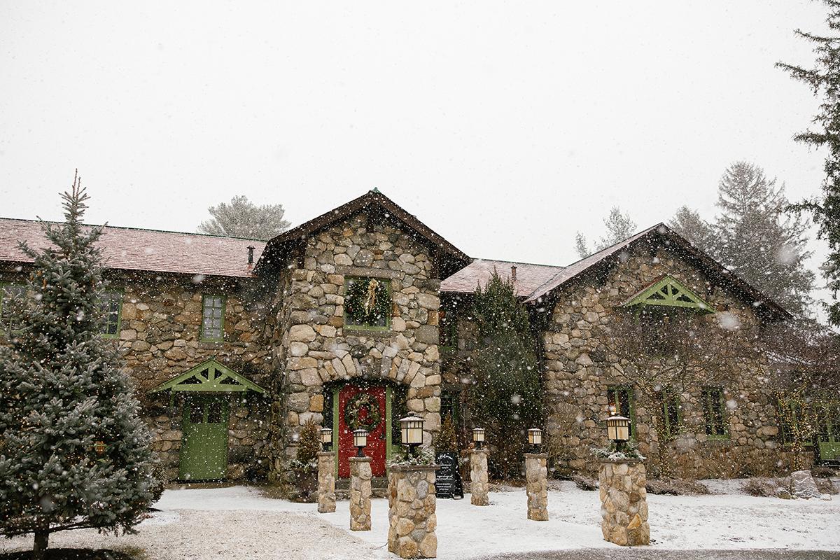 Snow-covered stone house with green doors and wreaths, framed by evergreens.