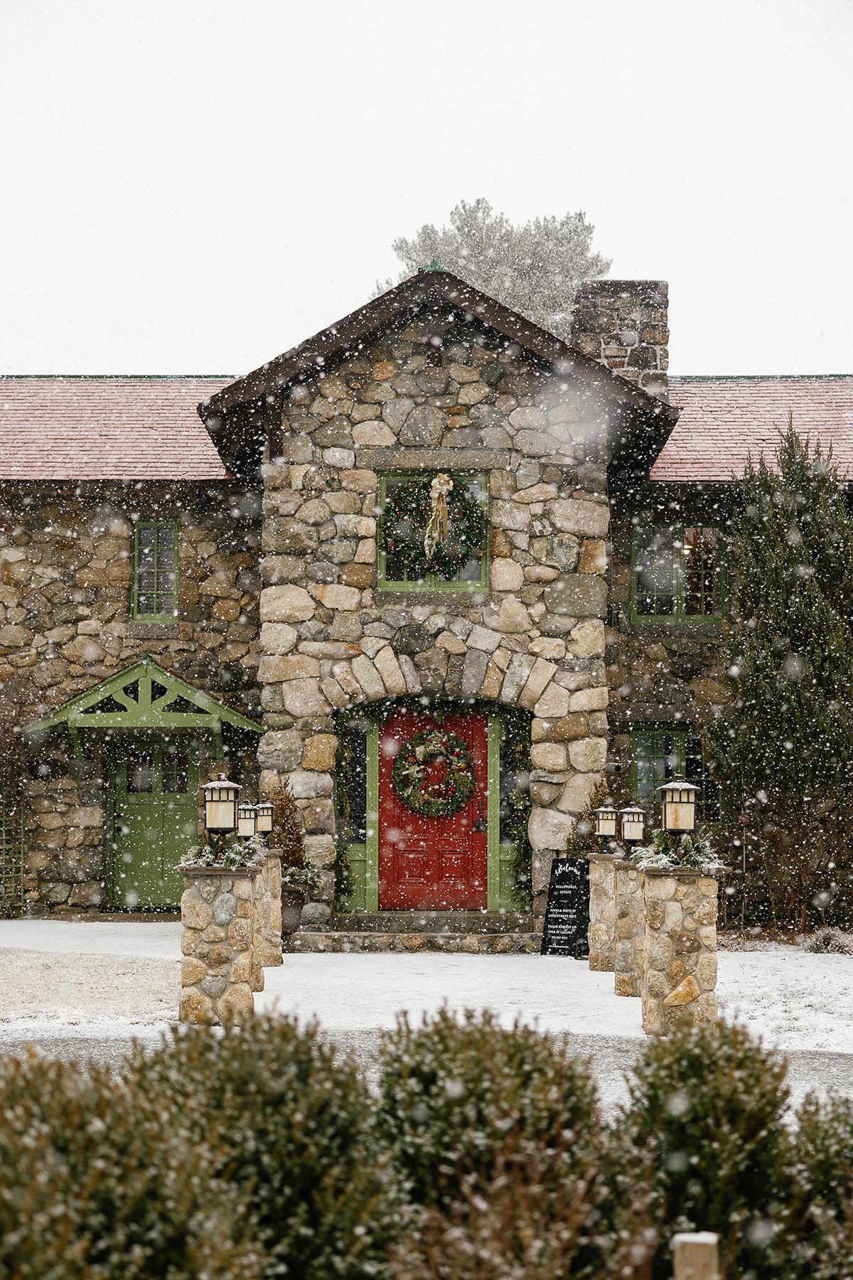 Snow-covered stone cottage with a red wreath-adorned door and evergreen shrubs