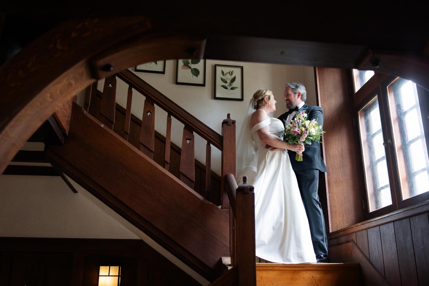 Bride and groom embrace on a wooden staircase by a sunlit window.
