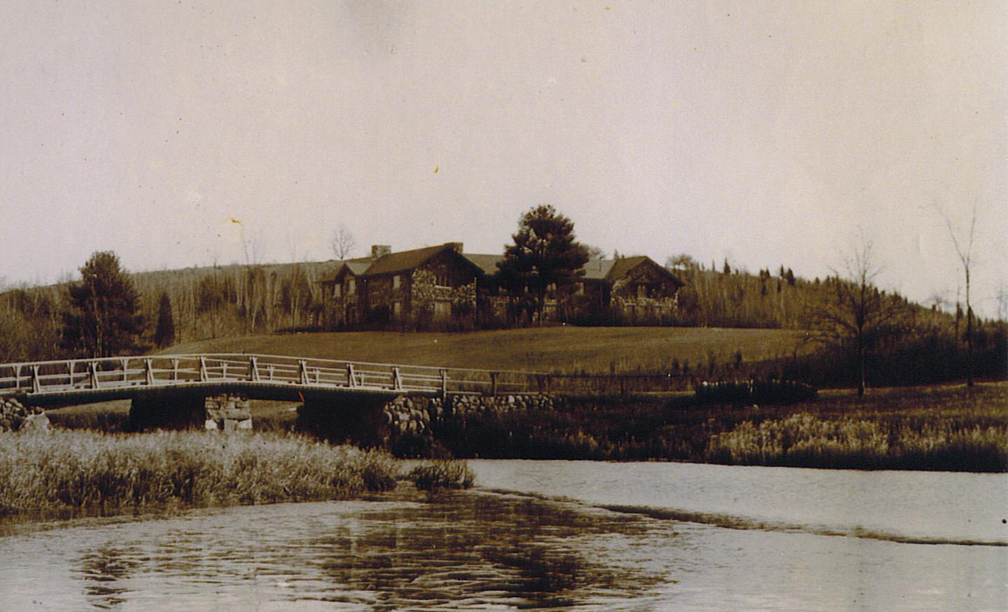 Sepia photograph of a rural house and bridge beside a calm river.