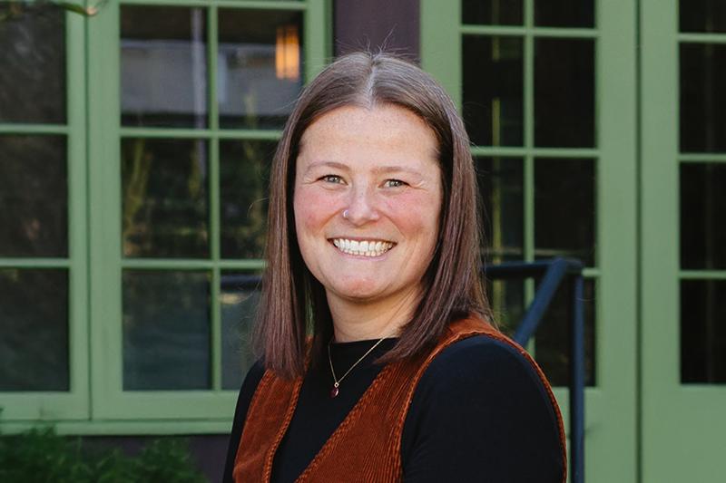 Smiling woman in a rust vest standing before green-paned windows.