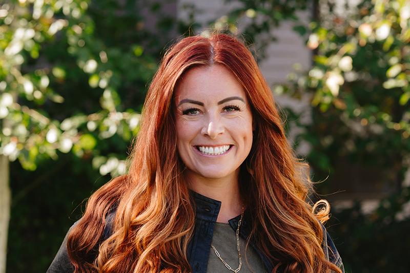 Smiling red-haired woman outdoors with sunlit greenery behind her.