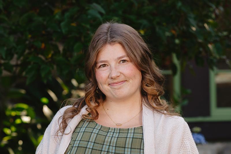 Smiling woman with wavy brown hair outdoors, wearing a plaid dress and light cardigan.